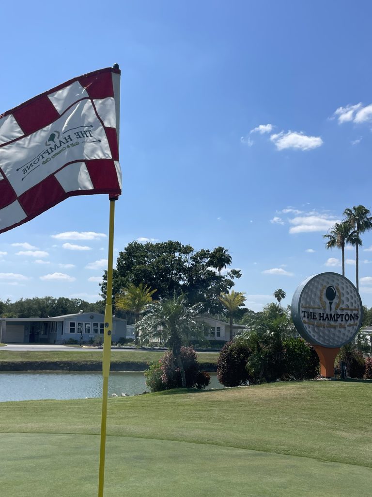 Golf course flag and Hamptons sign beside a pond on a sunny day.