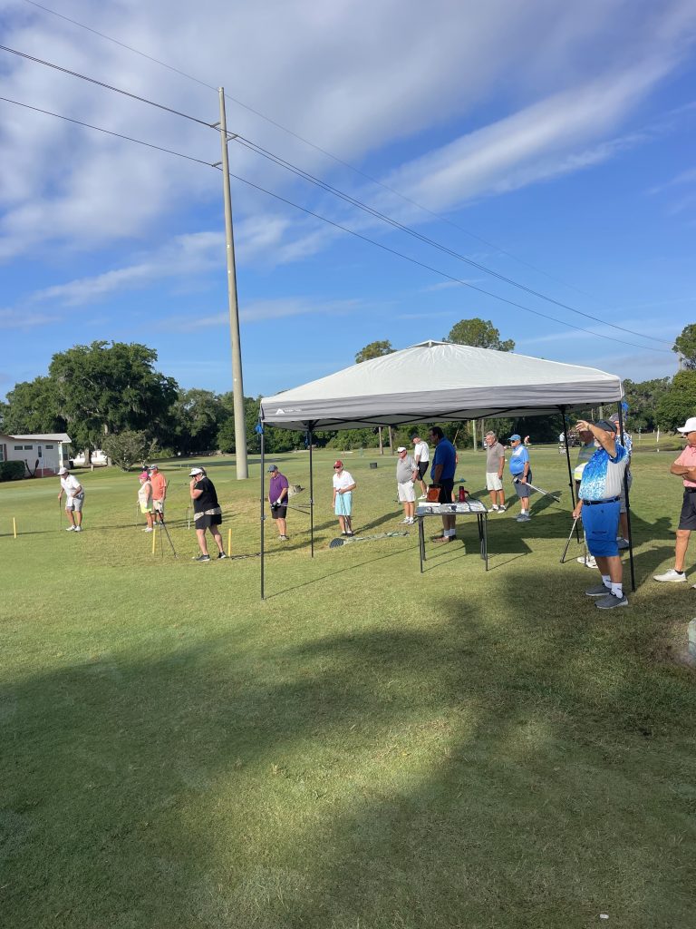 People gather under a canopy at a golf course during a sunny outdoor event.